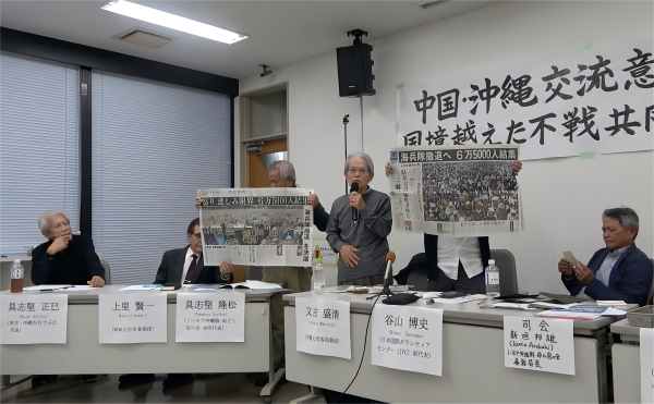 Professor Seikiyo Matayoshi displaying front-page reports from two 2016 newspapers: "Anger and Sorrow Reach a Breaking Point, 65,000 Gather in Protest" and "Mourning the Victim and Demanding the Withdrawal of the Marine Corps, 65,000 Participate in Okinawa Prefectural Assembly."